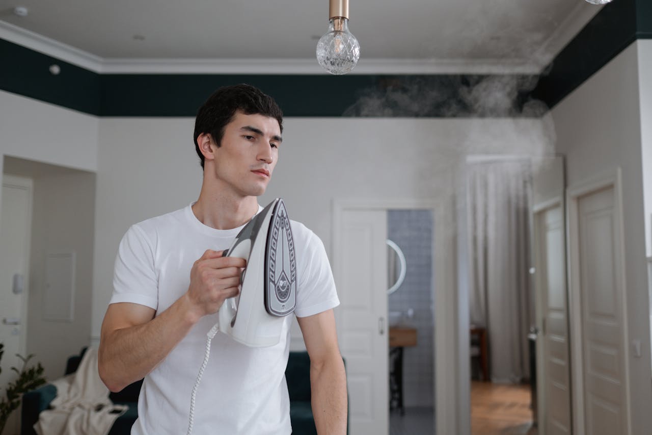 A man uses a steam iron on a white shirt indoors, highlighting daily household chores.